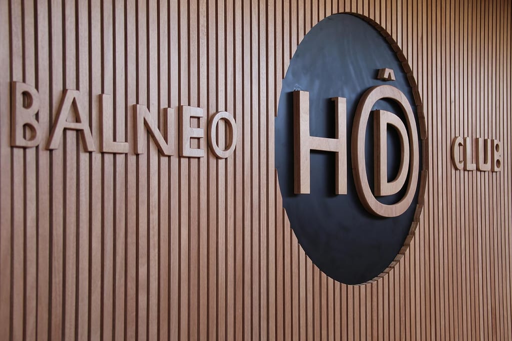 Enseigne de façade en bois avec “BALNÉO” et “CLUB”, ainsi qu’une grande forme “HO”, éclairée doucement sur fond de lames verticales.