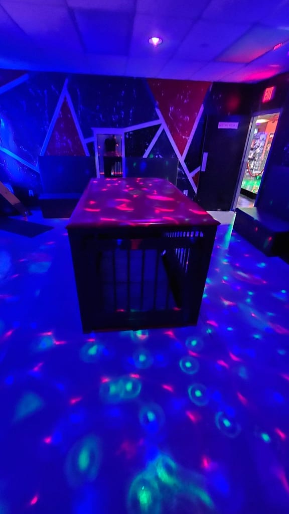 Violett und blau laserbeleuchteter Playroom mit gepolsterter Plattform in der Mitte, geometrischer Wanddekoration und farbigen Disco-Lichtreflexen auf dem Boden.