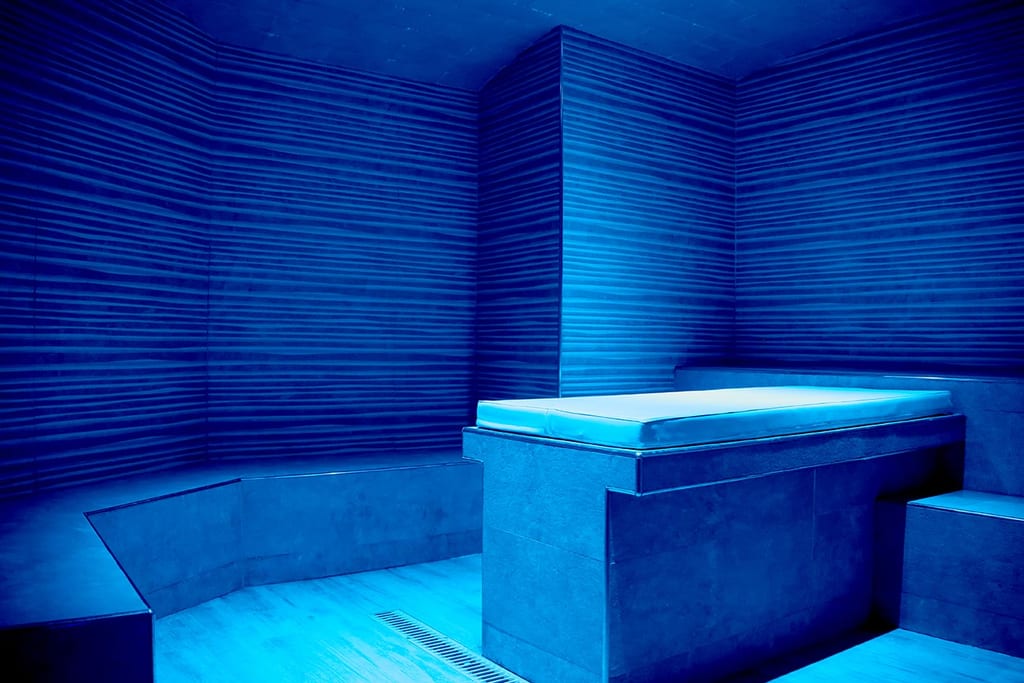Design de hammam em pedra e madeira iluminado a azul, com bancos e um ambiente de spa silencioso e acolhedor.