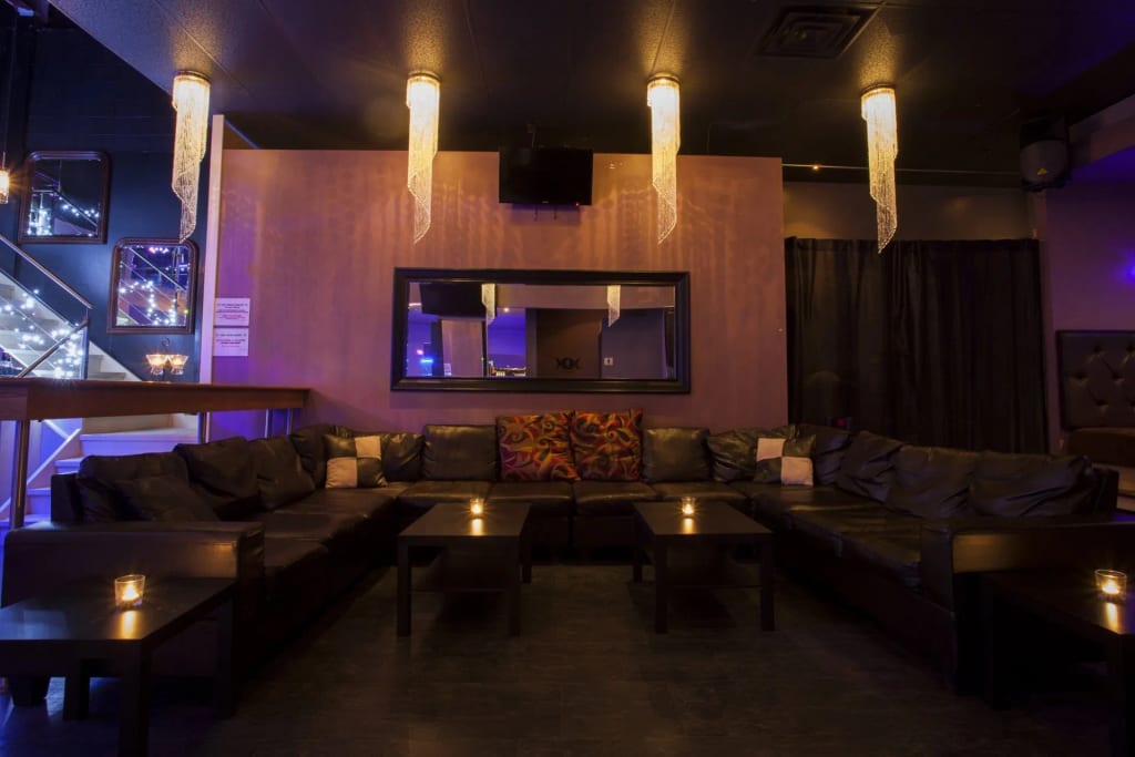 A dim lounge with plush dark sofas, candlelit tables, hanging crystal lights, and purple-blue ambiance lighting, showcasing the cozy @wishpride venue style.