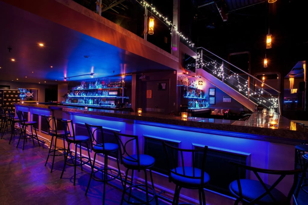 Um interior de bar moderno com um longo balcão iluminado, muitas garrafas de bebidas, iluminação de destaque azul e luminárias pendentes acesas, em sintonia com a atmosfera @wishpride.