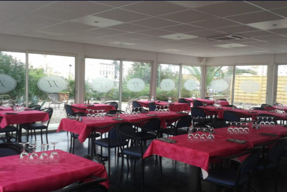 Salle à manger avec nappes rouges, assiettes et couverts soigneusement dressés, et grandes baies vitrées sous un plafond moderne et lumineux.