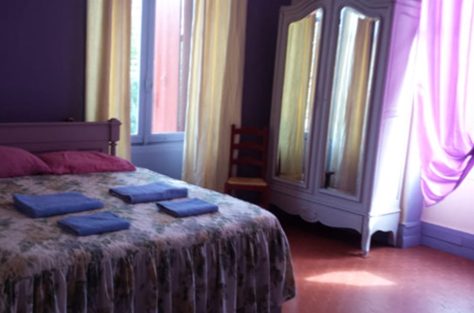 Chambre cosy avec lit fait, rideaux superposés aux tons doux et lumière chaude teintée de violet près des fenêtres.