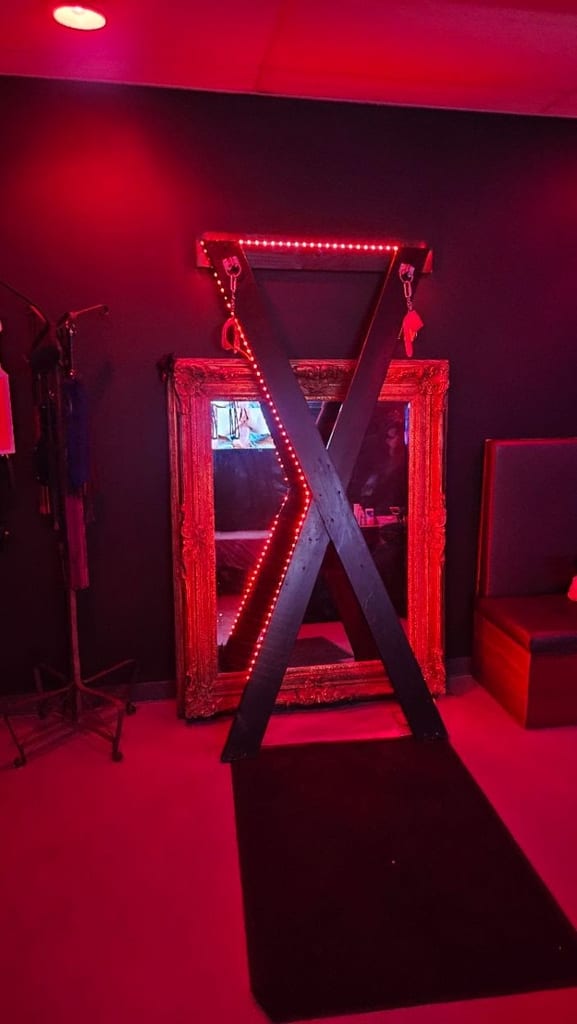Escena de playroom iluminada en rojo neón con una estructura en X, un panel de espejo, correas colgantes y un sofá frente a paredes oscuras bajo una suave luz de techo.