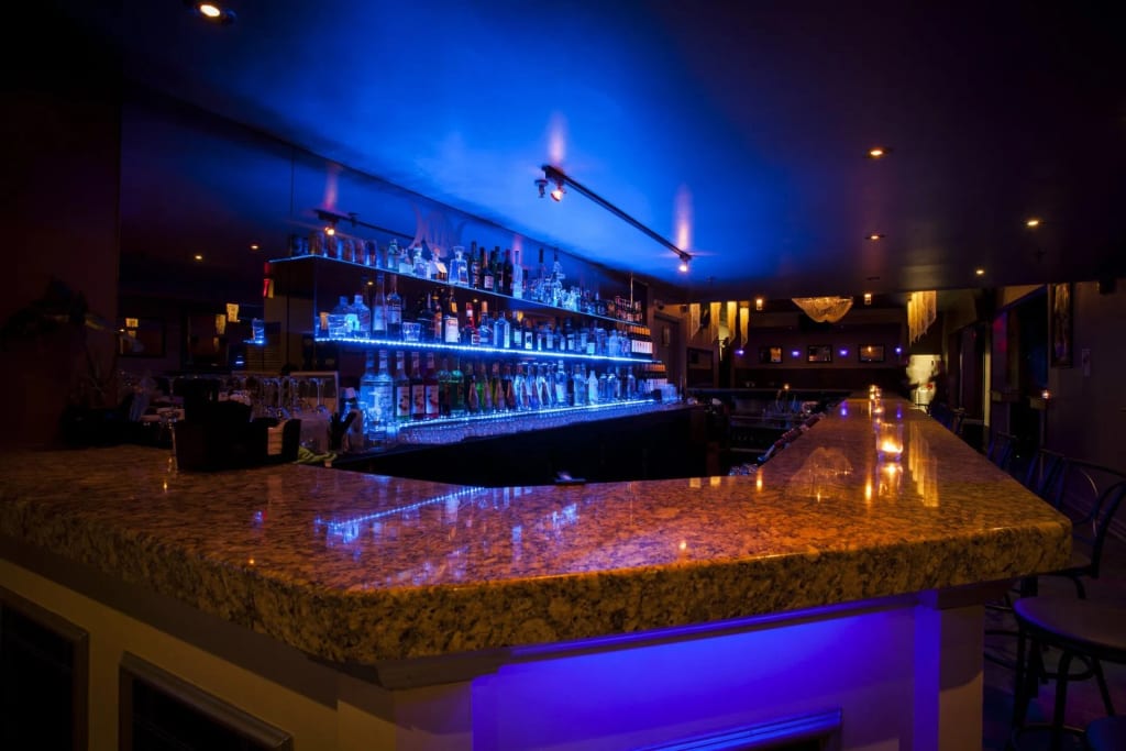 Un’area bar di alto livello con un lungo bancone, scaffali luminosi per bottiglie a LED blu e lampade a sospensione calde sotto un soffitto scuro, nello stile @wishpride.