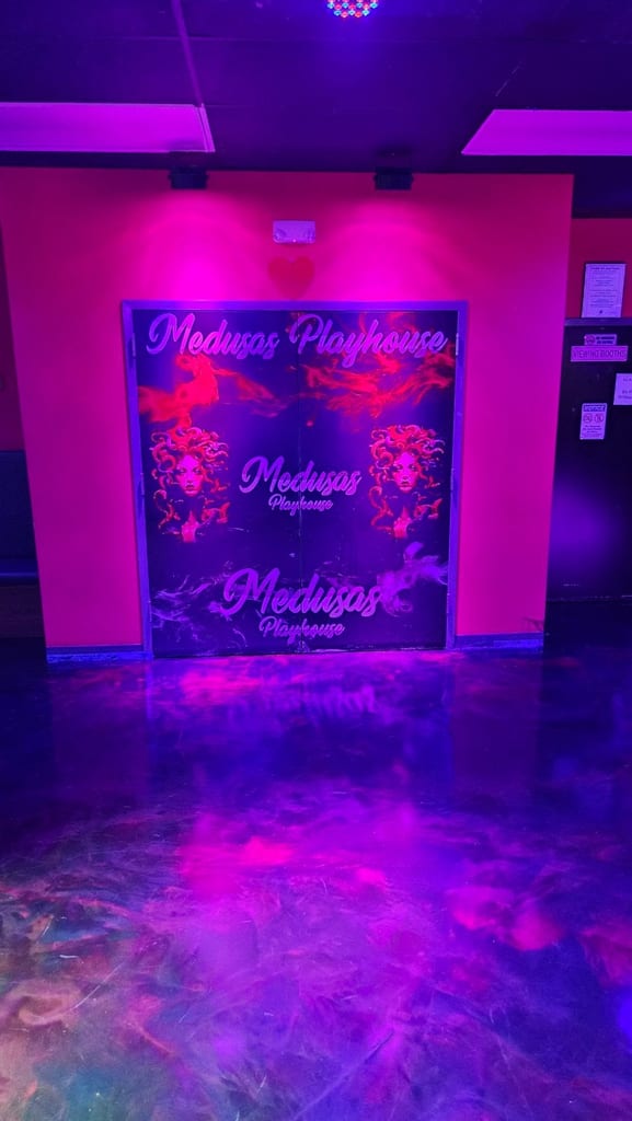 Salle violet néon avec une porte vitrée portant l’inscription « Medusa’s Playhouse », un éclairage lounge au-dessus et des panneaux d’information le long du mur.