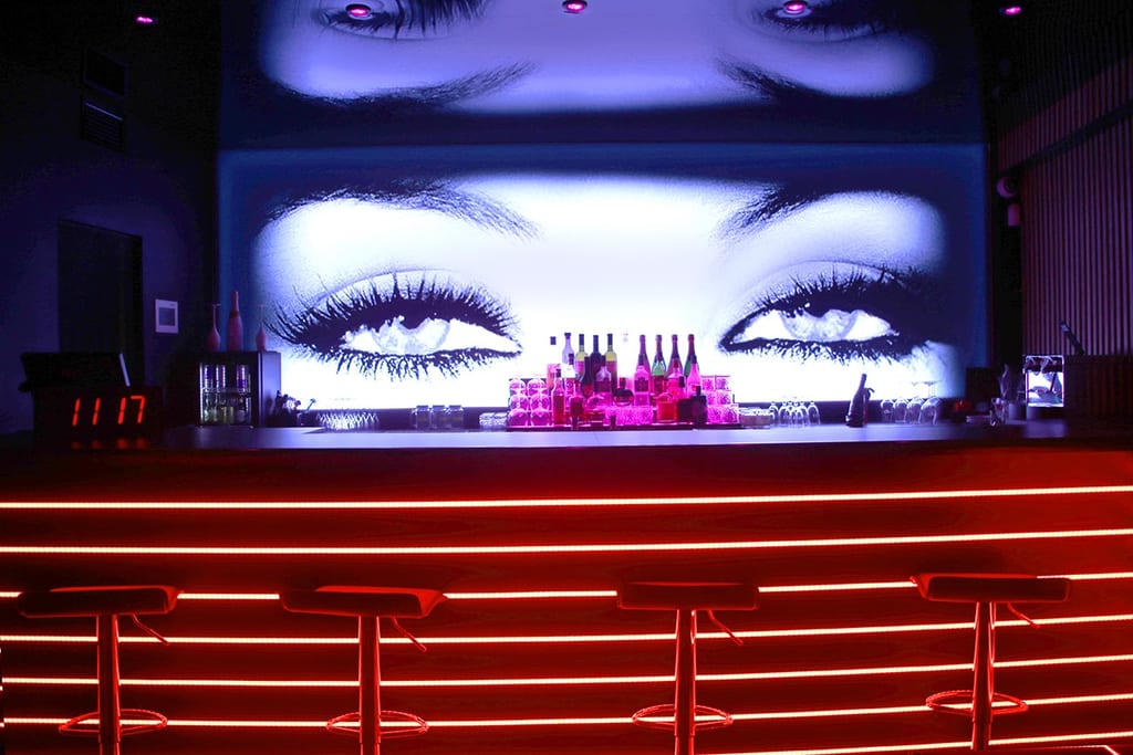 A neon bar counter with red LED strips, colorful bottles and a large eye artwork projected above it for a nightlife vibe.