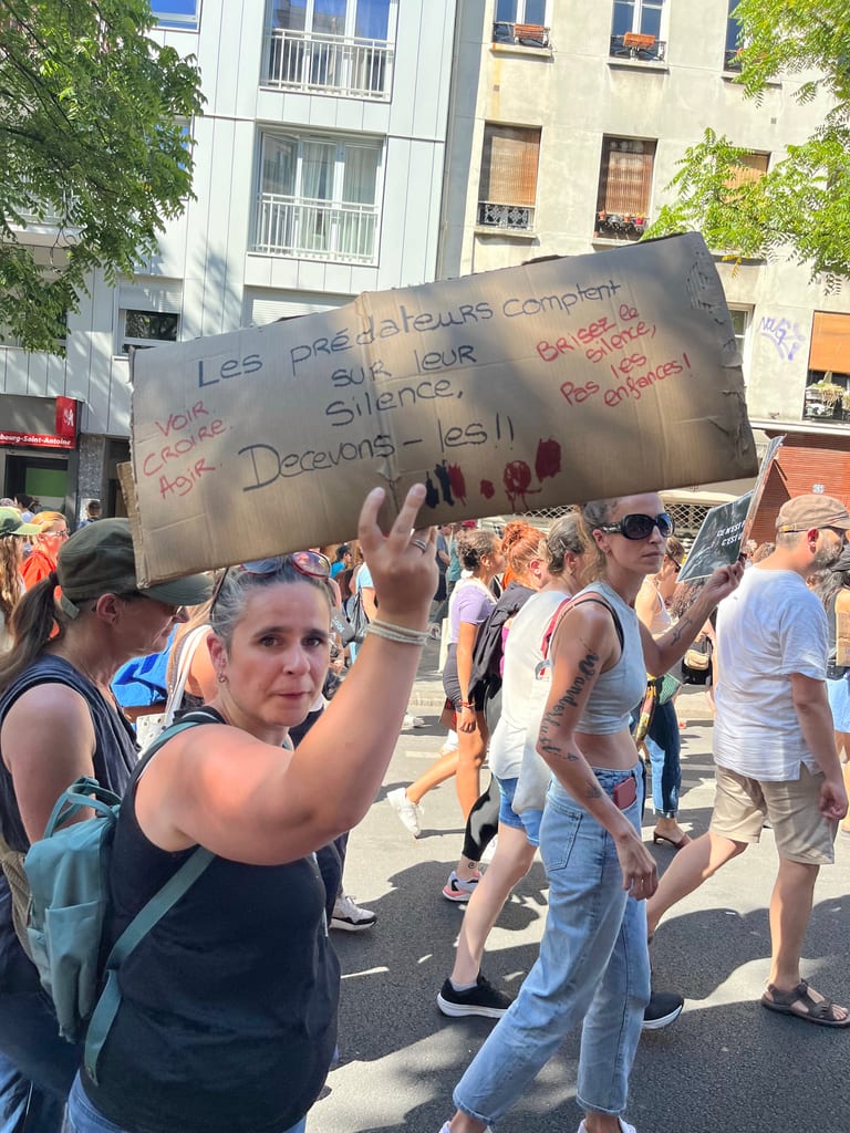 Crowd at a demonstration holding a cardboard sign about predators, silence, and “Say nothing” in bright colors.