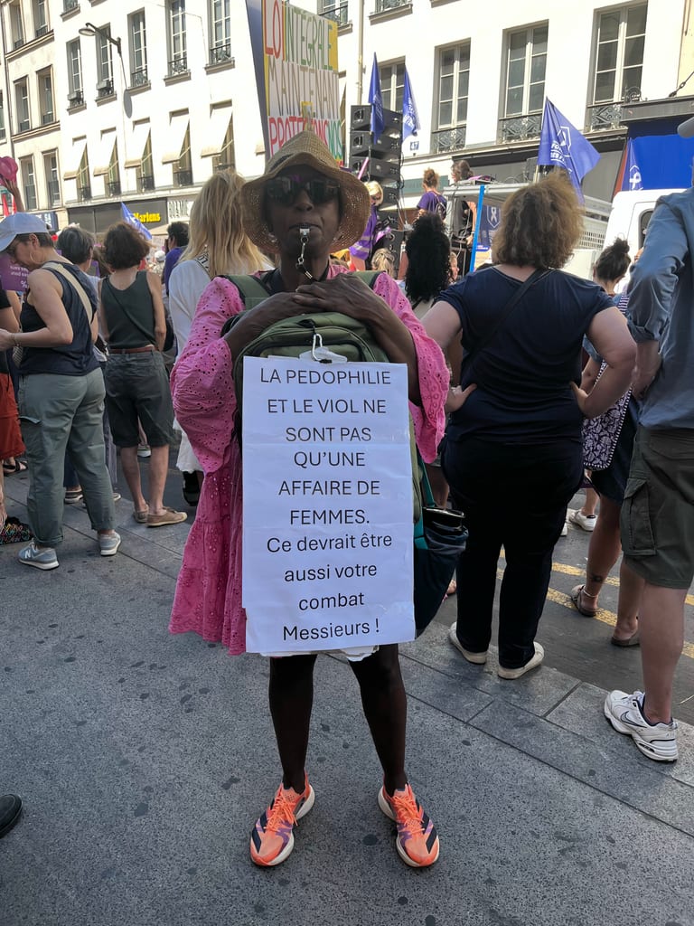 Protester in a straw hat holding a large placard about pedophilia and violence during a street rally with flags and tents behind, photo by @Floe.