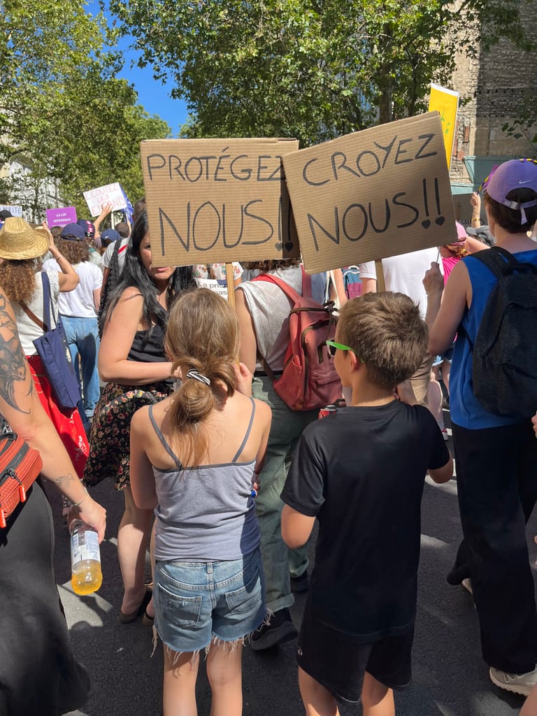 Des manifestants dans une rue ensoleillée portent de grandes pancartes en carton disant « PROTÉGEZ… » et « NOUS !! » au milieu d'une foule dense.