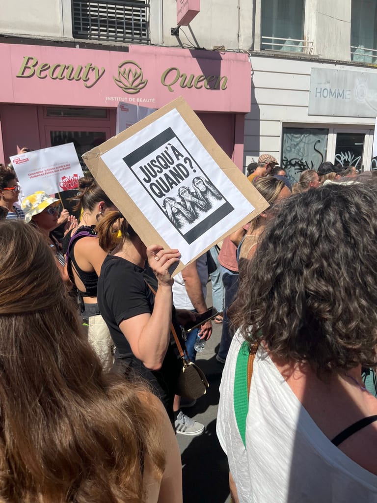 A protest poster reading “How much longer?” held in front of the “Beauty Queen” storefront, with a crowd behind it.